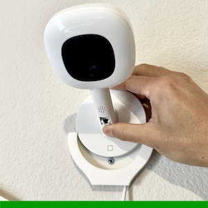 May include: A white security camera with a round base is being installed on a white wall. The camera is being held by a hand and is being rotated to adjust the angle. The camera has a black lens and a small microphone on the side. The base of the camera has a small screw hole and a small square hole. The camera is easy to insert and remove.