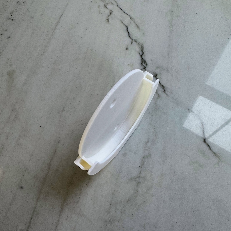May include: White plastic wall mount with two holes for hanging. The mount is designed to hold a round object, such as a clock or a mirror.