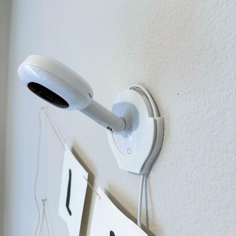 May include: A white baby monitor with a round base and a swiveling camera lens. The monitor is mounted on a white wall.