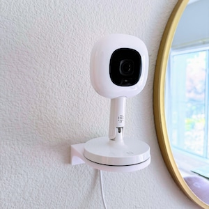 May include: A white baby monitor with a round camera lens and a white base. The monitor is mounted on a white wall with a white cord.