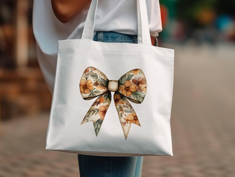 May include: A white tote bag with a floral bow design. The bow is made of watercolor flowers in shades of orange, yellow, and brown.