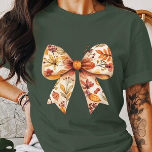 May include: A green t-shirt with a floral bow design. The bow is made of white fabric with orange and brown floral accents.