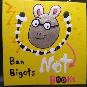 May include: A yellow canvas art piece featuring a cartoon aardvark wearing glasses. The text "Ban Bigots Not Books" is written on the canvas. The aardvark is surrounded by a black and white design and a yellow lightning bolt.