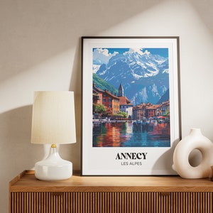 May include: A framed print of a colorful painting depicting a village nestled in the French Alps. The painting features a lake with boats, a mountain range in the background, and the text "Annecy Les Alpes".
