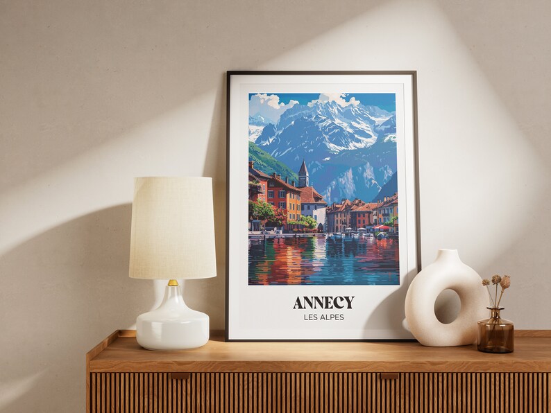May include: A framed print of a colorful painting depicting a village nestled in the French Alps. The painting features a lake with boats, a mountain range in the background, and the text "Annecy Les Alpes".