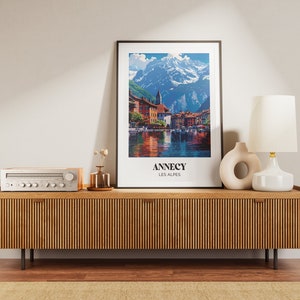 May include: A wooden media console with a light wood finish and vertical grooves. A vintage stereo receiver sits on top of the console. A framed poster with a mountain scene and the text "Annecy Les Alpes" is on the wall above the console. A white ceramic lamp with a white shade and a ceramic vase with a circular opening are on the console.