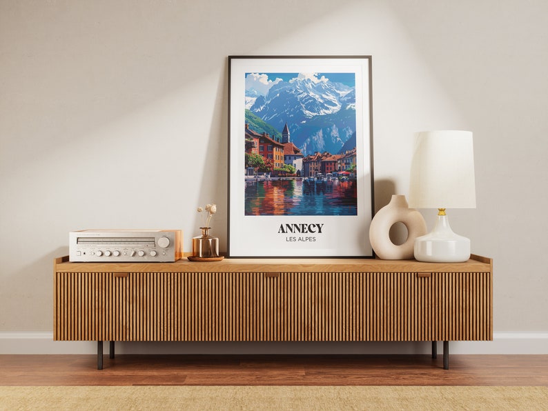 May include: A wooden media console with a light wood finish and vertical grooves. A vintage stereo receiver sits on top of the console. A framed poster with a mountain scene and the text "Annecy Les Alpes" is on the wall above the console. A white ceramic lamp with a white shade and a ceramic vase with a circular opening are on the console.