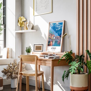 May include: A home office workspace with a wooden desk, a chair, and a framed print of a mountain scene. The desk has a lamp, a potted plant, and a few books on it. There is a large window with a view of a city skyline. The walls are white and the floor is wooden.