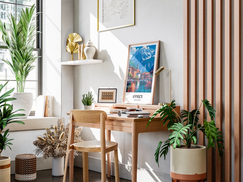 May include: A home office workspace with a wooden desk, a chair, and a framed print of a mountain scene. The desk has a lamp, a potted plant, and a few books on it. There is a large window with a view of a city skyline. The walls are white and the floor is wooden.