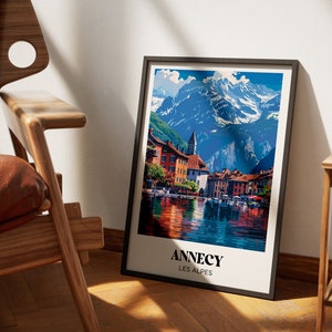 May include: A framed poster with a colorful illustration of a town by a lake with mountains in the background. The poster has the text "Annecy Les Alpes" at the bottom.