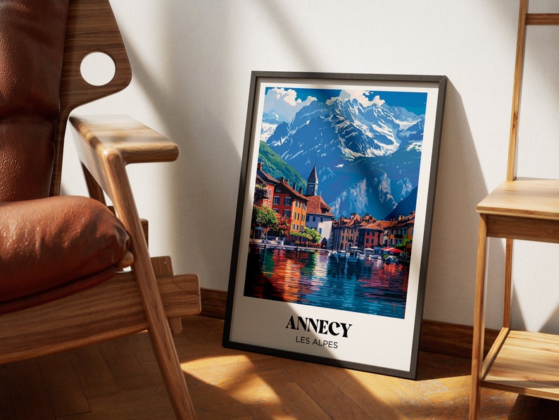 May include: A framed poster with a colorful illustration of a town by a lake with mountains in the background. The poster has the text "Annecy Les Alpes" at the bottom.