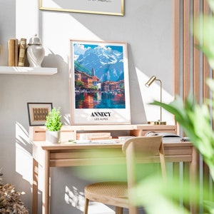 May include: A framed poster with a colorful illustration of a town on a lake with mountains in the background. The poster has the text "Annecy Les Alpes" at the bottom.