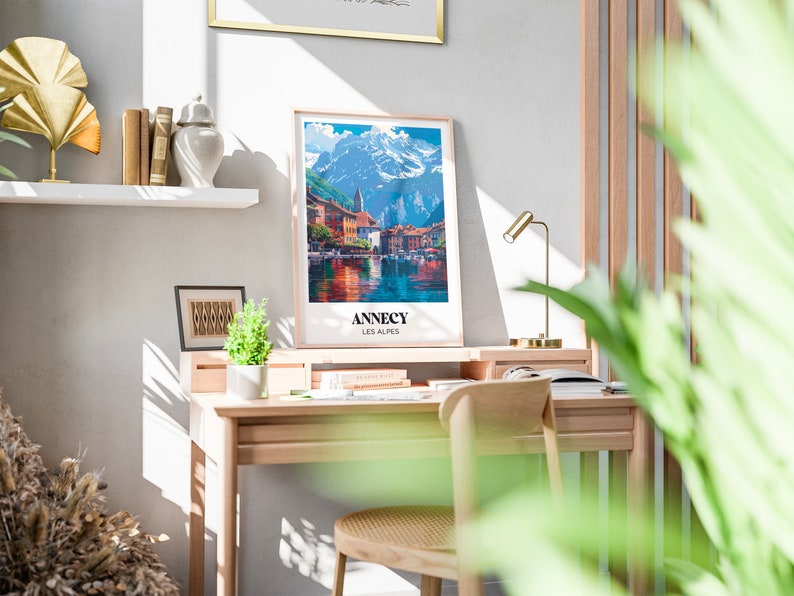 May include: A framed poster with a colorful illustration of a town on a lake with mountains in the background. The poster has the text "Annecy Les Alpes" at the bottom.