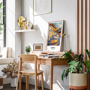 May include: A home office workspace with a wooden desk, chair, and a potted plant. The desk has a lamp, a framed print of a tram in Lisbon, Portugal, and a few books. There is a potted plant on the floor next to the desk.