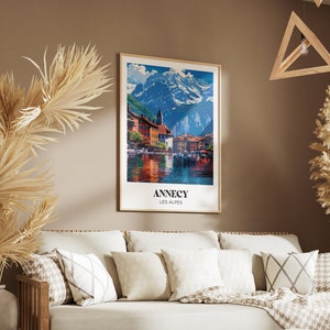 May include: A vintage travel poster featuring a scenic view of a town in the French Alps. The poster is framed in a light wood frame and hangs on a brown wall. The poster reads "Annecy Les Alpes".
