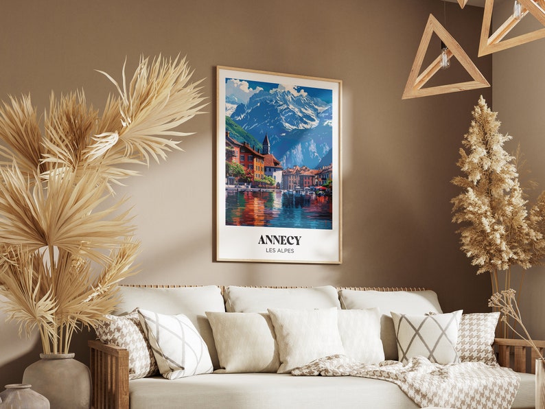 May include: A vintage travel poster featuring a scenic view of a town in the French Alps. The poster is framed in a light wood frame and hangs on a brown wall. The poster reads "Annecy Les Alpes".