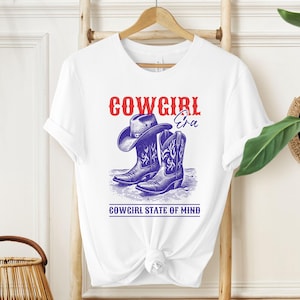 Puede incluir: Camiseta blanca con un gráfico azul y rojo de un sombrero y botas de vaquero. El texto "COWGIRL" es rojo, "Era" está en azul y "COWGIRL STATE OF MIND" está debajo. La camiseta está colgada en una percha de madera.