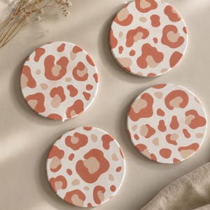 Könnte beinhalten: Ein Set aus drei runden Untersetzern mit einem sich wiederholenden Leopardenmuster in Terrakotta, Creme und Hellbeige. Jeder Untersetzer hat eine glatte, flache Oberfläche mit abgerundeten Kanten und zeigt ein einheitliches Tiermuster.