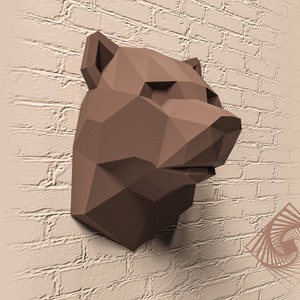 Może przedstawiać: Brązowa rzeźba papierowa głowy niedźwiedzia, z geometrycznym wzorem w stylu origami. Rzeźba jest zamontowana na białej ceglanej ścianie.