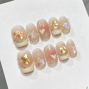 May include: A set of ten nude-coloured press-on nails with gold leaf accents and small white hearts. The nails are displayed on a white card with the text "Nail Studio Make It Romantic And Meaningful".