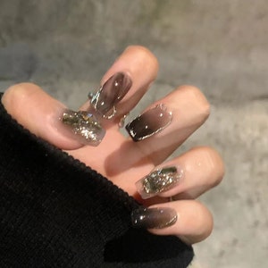 May include: Close-up of a hand with long, square-shaped nails. The nails are painted with a gradient of black and brown, and some have silver glitter accents.