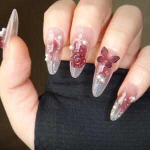 May include: Close-up of a hand with long, clear, almond-shaped nails decorated with red roses, butterflies, and silver glitter.