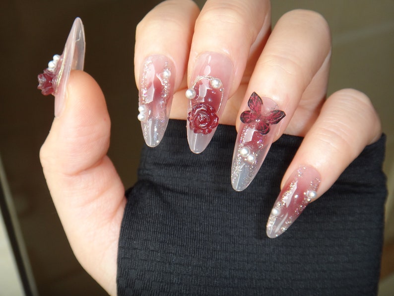 May include: Close-up of a hand with long, clear, almond-shaped nails decorated with red roses, butterflies, and silver glitter.
