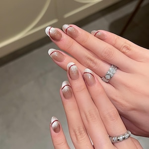 Puede incluir: Diseño de uñas ovaladas con un efecto degradado de una base nude a una punta blanca brillante. Una fina línea roja acentúa las uñas. Se muestran dos manos, cada una con un anillo plateado.