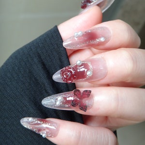May include: Close-up of a hand with long, clear, oval-shaped acrylic nails decorated with red roses, silver glitter, and small white pearls.