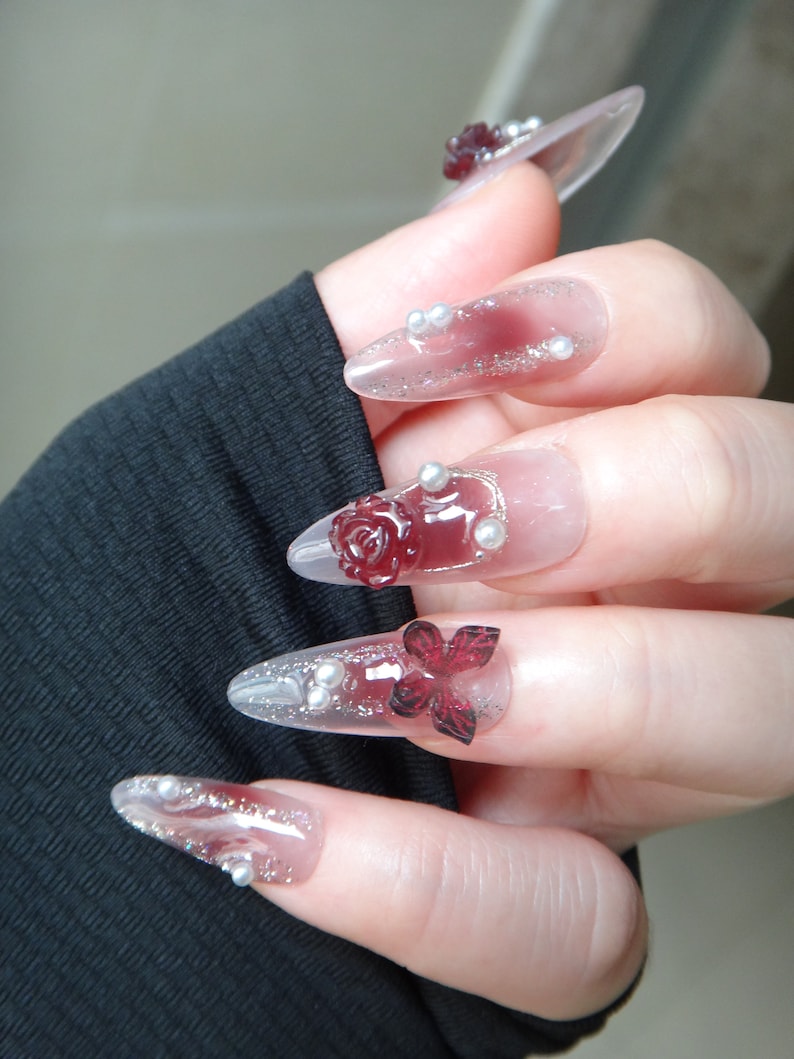 May include: Close-up of a hand with long, clear, oval-shaped acrylic nails decorated with red roses, silver glitter, and small white pearls.