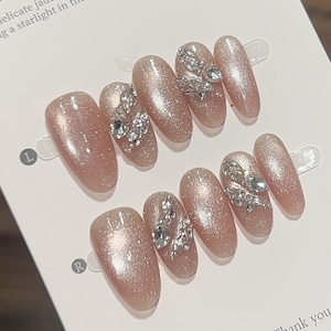 Peut inclure: Ensemble de dix faux ongles de couleur nude avec des paillettes argentées et des strass. Les ongles sont disposés en deux rangées, étiquetées "L" et "R".