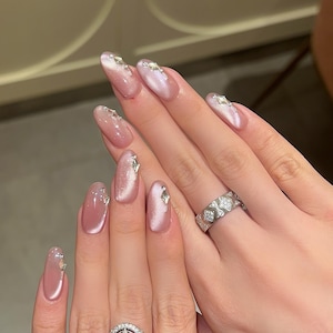 May include: Close-up of hands with oval-shaped nails painted a light pink with a shimmery finish. Each nail is adorned with small, clear rhinestones. A silver ring with a diamond-like design is worn on the ring finger of the right hand.