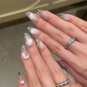 以下が含まれることがあります： 銀色と白の虹色の光沢のあるポリッシュで塗られた、アーモンド型の長い爪をした手のクローズアップ。各爪には小さな銀色のアクセントと透明なラインストーンが施されています。