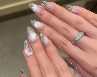 May include: Close-up of hands with long, almond-shaped nails painted with a silver and white iridescent polish. Each nail has a small silver accent and a clear rhinestone.