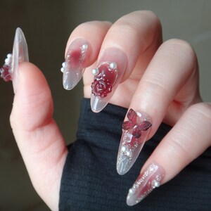 May include: Close-up of a hand with long, clear, almond-shaped nails decorated with red and white 3D flowers and butterflies. The nails are also adorned with small white pearls.