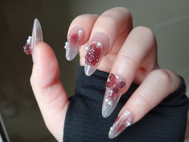 May include: Close-up of a hand with long, clear, almond-shaped nails decorated with red and white 3D flowers and butterflies. The nails are also adorned with small white pearls.