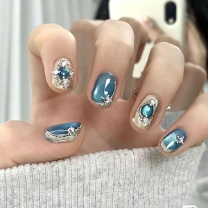 May include: Close-up of manicured nails with a blue and silver design. The nails feature a combination of metallic silver and glossy blue polish, adorned with jewel-like embellishments. The design includes oval and rectangular shapes, creating a glamorous and elegant look.