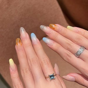 May include: Close-up of manicured nails with a gradient design in shades of yellow, orange, blue, and clear glitter. The nails are long and oval-shaped. Two hands are shown, one with a gold ring with a square blue stone and another with a silver band.