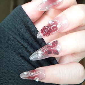 May include: Close-up of a hand with long, clear, pointed nails decorated with red roses, glitter, and silver beads.