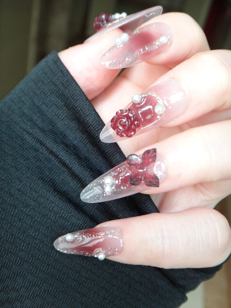 May include: Close-up of a hand with long, clear, pointed nails decorated with red roses, glitter, and silver beads.