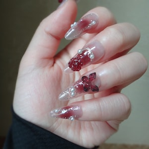 May include: Close-up of a hand with long, pointed, clear acrylic nails decorated with red and white 3D flowers and pearl accents.