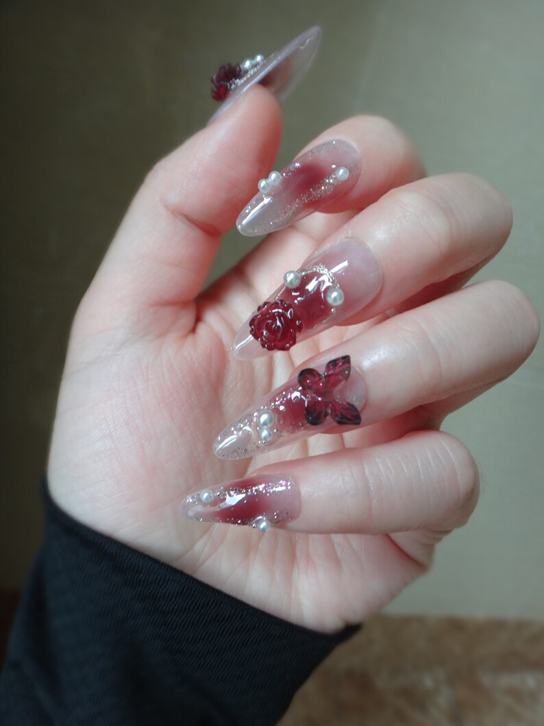 May include: Close-up of a hand with long, pointed, clear acrylic nails decorated with red and white 3D flowers and pearl accents.