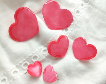 May include: Five pink heart-shaped resin ornaments of varying sizes. The hearts are on a white fabric background.