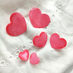 Puede incluir: Cinco adornos de resina en forma de corazón rosa de diferentes tamaños. Los corazones están sobre un fondo de tela blanca.