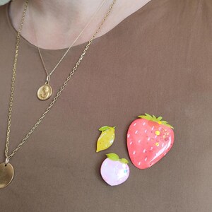 Peut inclure: Ensemble de broches en forme de fruits : une fraise rouge avec des feuilles vertes, un citron jaune et une pêche rose. Les broches sont posées sur un haut marron, avec des colliers dorés en arrière-plan.