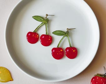 May include: A pair of cherry earrings with red, glossy cherries and green stems and leaves, displayed on a white plate. Other fruit-shaped earrings, including a strawberry, peach, and lemon, are scattered around the plate.