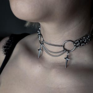 May include: A silver-toned metal choker necklace featuring a chainmail band connected to two circular rings. Three delicate draped chains hang between the rings, and two four-pointed star-shaped charms dangle from the bottom of each ring.