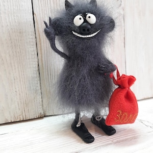 May include: A gray, furry, cartoon-like creature with a big smile and large eyes, holding a red bag with gold lettering. The creature is standing on a white wooden surface.
