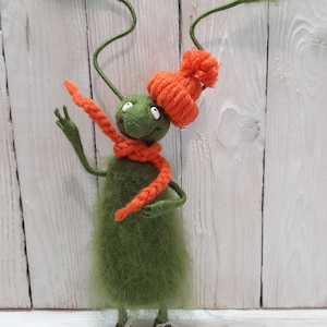 Könnte beinhalten: Ein grünes Filzspielzeug mit langen, dünnen Armen und Beinen, das eine orangefarbene Strickmütze und einen Schal trägt. Das Spielzeug hat große, weiße Augen und einen freundlichen Ausdruck.