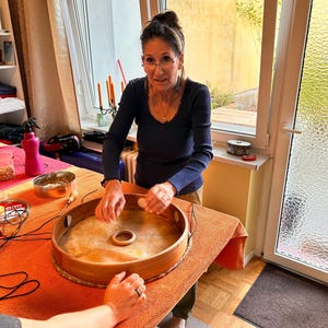 Puede incluir: Un tablero de juego de madera con una superficie de juego circular y un agujero central. El tablero está sobre un mantel naranja. Una persona sostiene una pieza de juego blanca, preparándose para jugar. El juego es probablemente un juego de mesa.